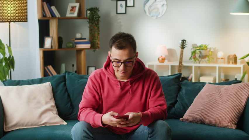 Asian man using smartphone. Young Korean guy texting at phone, sitting on sofa in living room, male person relaxing at home. Communicating with family and friends.  Royalty-Free Stock Footage #1088194915