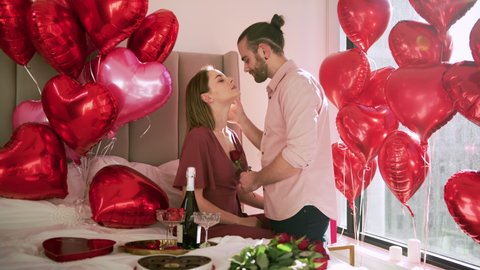 Sensual couple embracing and kissing in bed slow motion. Passionate man and woman enjoying sensual foreplay in decorated hotel room. Sexy lovers kissing in bedroom full of red heart balloons and roses