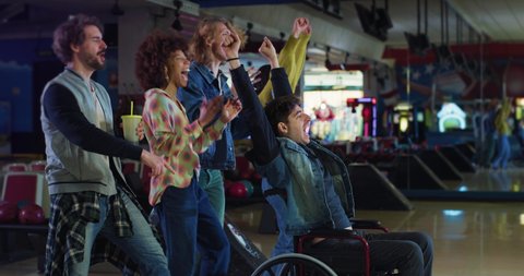 Cinematic shot of young man with disability who uses wheelchair throwing strike ball and exulting victory while having fun to play bowling together with multiracial college friends in sporting club.