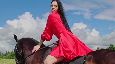 Beautiful young woman riding a horse on field, wearing fashionable red dress. Girl horseback caresses dark brown horse