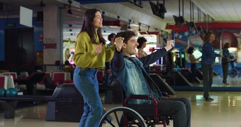 Cinematic shot of young man with disability who uses wheelchair is throwing strike ball and exulting victory while having fun to play bowling together with his girlfriend on weekend in sporting club.