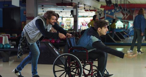 Cinematic shot of young man with disability who uses wheelchair throwing strike ball and exulting victory while having fun to play bowling together with his college friend on weekend in sporting club.