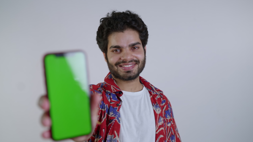 Young Indian Asian Smiling male extending hand showing Green Screen or Chroma key on a Smartphone. A male or trendy stylish guy with a new mobile phone app or a brand isolated on white background.  Royalty-Free Stock Footage #1088200105