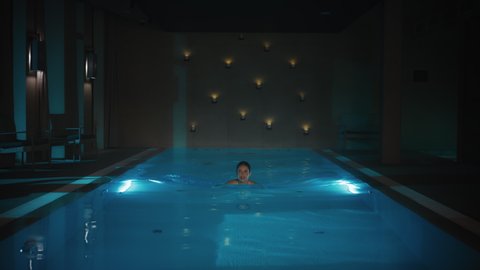 Still Shot of a Beautiful Caucasian Female Swimming in a Blue Light Pool with Cangles Burning in the Background. Gorgeous Brunette Woman Taking a Swim at a Hotel Spa.