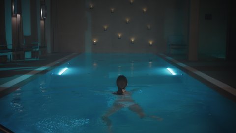 Beautiful Caucasian Female Swimming in a Blue Light Pool with Cangles Burning in the Background. Gorgeous Brunette Woman Taking a Swim at a Hotel Spa.