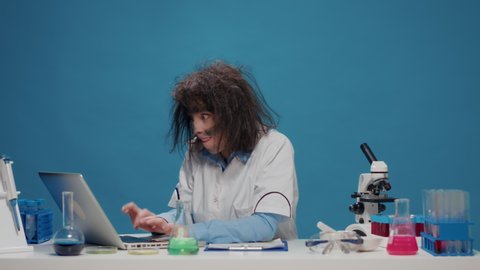 Insane foolish female chemist working with microscope in studio, acting crazy and insane on camera. Goofy amusing woman feeling silly and being funny, mad playful scientist with messy hair.