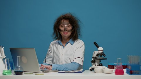 Portrait of crazy female chemist working on laptop at desk, being funny and goofy in studio. Mad insane scientist using computer and doing foolish silly facial expressions, having messy hair.