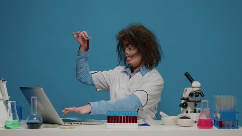 Funny insane scientist with messy hair looking at test tubes, feeling confident sitting over blue background. Mad goofy chemist acting crazy and foolish, being amusing and funny.