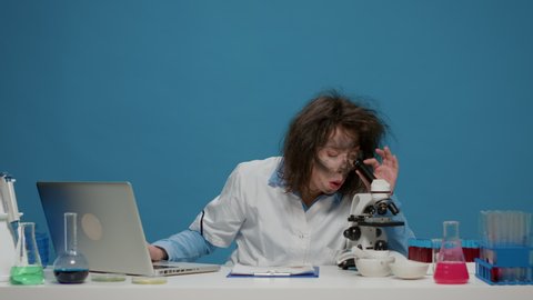Crazy foolish chemist using lab microscope over background, acting goofy and grinning. Mad amusing scientist with messy hair looking silly and funny, insane fun hairstyle in studio.