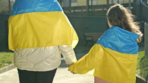 An adult woman holds the hand of a little girl. The shoulders of both are covered with the Flag of Ukraine. Back view.