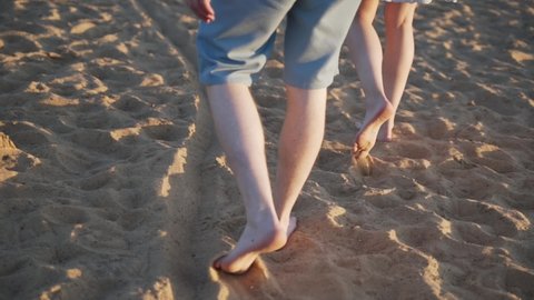 Male caucasian hairy legs in shorts and female legs in skirt walk barefoot on the sand in the setting sun, close-up. Couple in love walks along the dry sandy shore on summer evening.