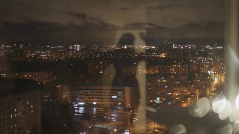 View from high floor of big city at night with high-rise buildings with lights on in the windows. In reflection, the silhouette of a unrecognizable woman takes off her clothes and leaves.