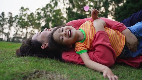 Lovely young woman laying on grass playing with little cute girl with a lollipop in hand, laughing happily, girl laying on mother’s belly, having fun together