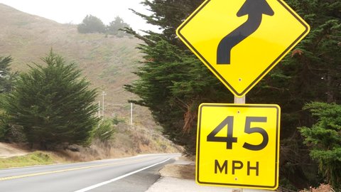 Speed limit sign, yellow warning or caution about curves, turns or winding road ahead. Pacific coast highway trip, cabrillo highway along ocean coast. Cars driving on asphalt. California Big Sur, USA.