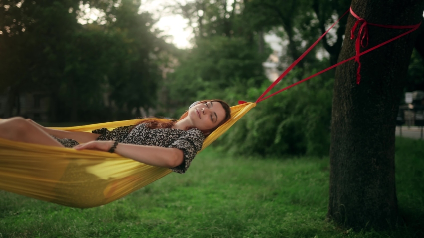 Young beautiful red-haired woman lying in a hammock. Attractive girl smiles while listening to music wearing wireless headphones