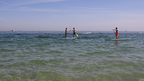Children ride SUP stand up paddle boards on sea coast, boats pass in background. Seascape of purest sea water, sandy seabed. Summer water sport standup paddleboarding. Odesa, Ukraine, 09 13 2020