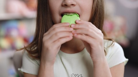 Close up front view, unrecognizable kid brunette girl with big smile and white healthy teeth bites pistachio macaron cookie. Tasty sweet color macaron