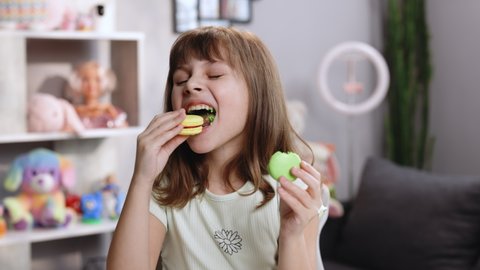 Attractive smiling cute school kid girl holds two tasty cookies in her hands and makes her choice, then bite macaron in yellow color and looking at camera. Traditional French multicolored macaroon