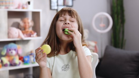 Young brunette kid girl can not make right decision and choice what macaron to bite first - left from with lemon and beryy filling or right made with pistachio cream, then bite macaron in green color