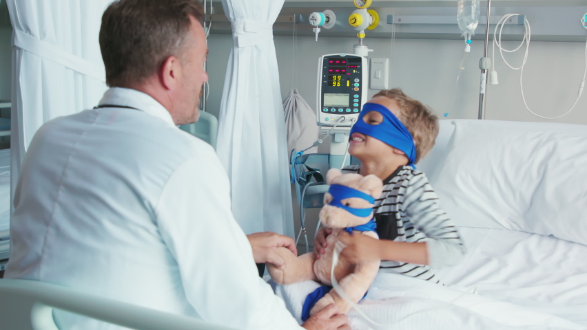 Cute little boy with superhero mask embracing teddy bear while playing with doctor. Playful child dressed as superhero holding stuff toy talking to pediatrician. Doctor visiting his little patient. Royalty-Free Stock Footage #1088219755