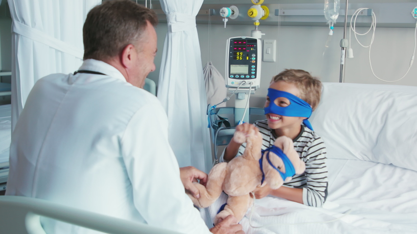 Cute little boy with superhero mask embracing teddy bear while playing with doctor. Playful child dressed as superhero holding stuff toy talking to pediatrician. Doctor visiting his little patient. | Shutterstock HD Video #1088219755