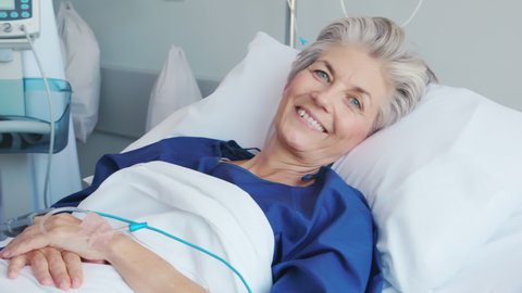 Mature woman recovering from illness while lying on hospital bed. Portrait of old hospitalized patient recovering after surgery. Happy senior woman in hospital ward while looking at camera.