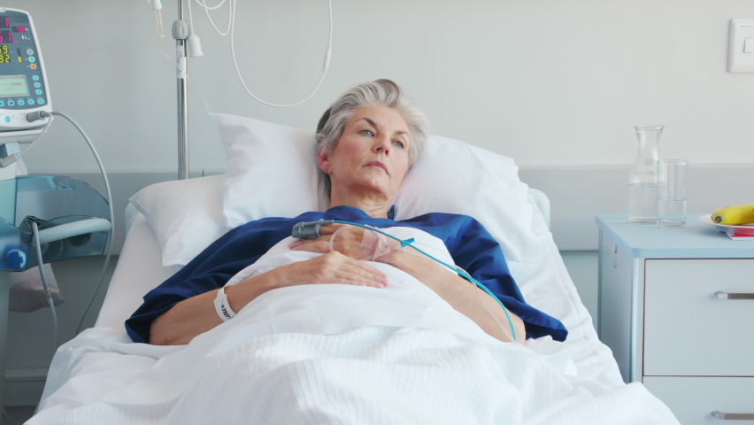 Pensive sad old woman lying on hospital bed looking trough the window. Sick lonely old patient under white blanket thinking on hospital bed. Mature thoughtful woman lying ill in bed in a clinic. Royalty-Free Stock Footage #1088219775