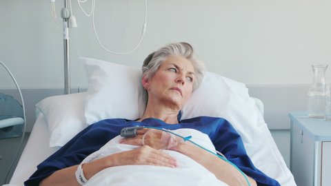 Pensive sad old woman lying on hospital bed looking trough the window. Sick lonely old patient under white blanket thinking on hospital bed. Mature thoughtful woman lying ill in bed in a clinic.