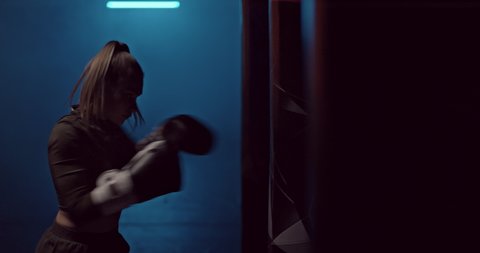 Silhouette, a woman boxer professionally makes strong punches on a punching bag in a dark hall of a fight club, preparing for a fight. Young female boxer practices punches in boxing gloves before a