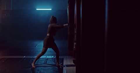 Female athlete boxing a punching bag in a dark hall of a fight club, punching bags hang in a row. Athlete professionally trains before the fight, practicing punches on a punching bag, silhouette. 4k
