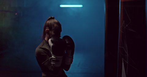Portrait, professional boxer girl hits a punching bag with gloves, dark gym with smoke. Beautiful boxer girl practices punches in boxing gloves, preparing for the competition. 4k, ProRes