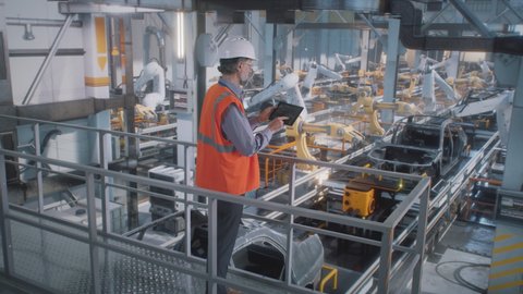 High angle of mature male engineer in uniform and hardhat using tablet to control robotic arms during work on contemporary car factory