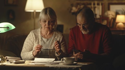 Elderly husband and wife analyzing utility bills and making calculations on smartphone while sitting near table in living room at home