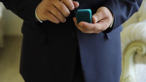 Groom in dark black classic suit opens blue box with wedding rings