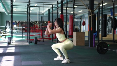 Wide shot portrait of confident Caucasian fit sportswoman squatting in gym indoors. Slim beautiful woman exercising working out in sports club. Athleticism and sport concept