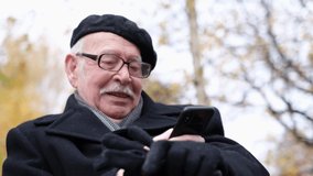 The pensioner uses a smartphone, he is sitting on a park bench. An 80-year-old pensioner