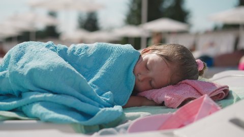 Tired little girl with wet hair covered with towel sleeps on deck chair on beach at marine resort on sunny summer day close view slow motion