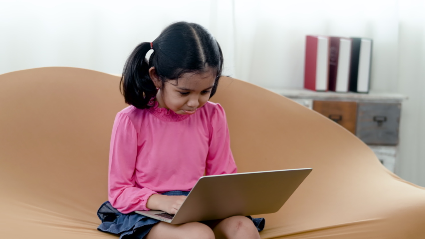 4K 50fps, Adolable Asian little girl wearing a pink shirt and blue skirt, sitting at her favorite brown sofa, sitting at her laptop computer, with a happy smiling face.	