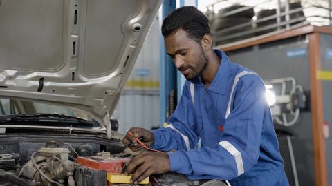 Car mechanic testing or checking car engine battery by using digital multimeter at garage - concept of technology, maintenance services and blue collar jobs.
