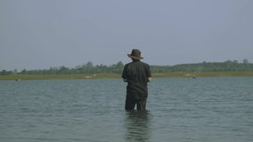 A man in a hat stands fishing in shallow water in the sunlight against a hazy sky background.