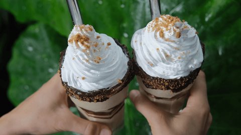 Two appetizing milkshakes with whipped cream with metal tubes for a cocktail are held in hands against the background of a green leaf of a tropical palm tree. Drinks on holiday at the hotel.