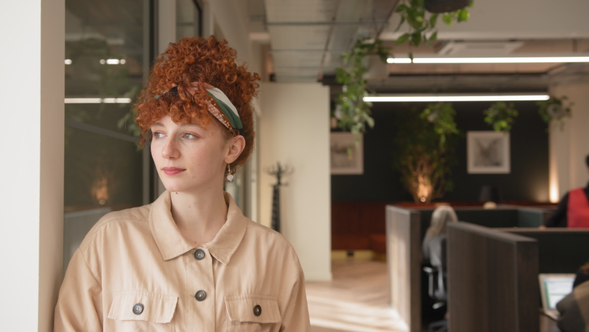 Slow motion of confident young white woman with red curly hair smiling towards camera in an open office space | Shutterstock HD Video #1088242971