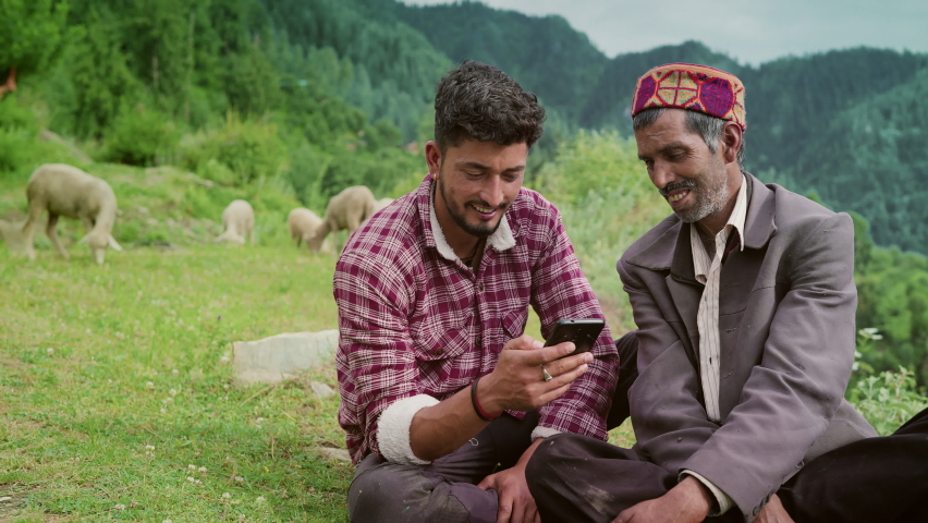 Smiling Indian elderly male shepherd with his young son sitting together using a Mobile phone with grazing sheep in a rural area of remote greenery mountainous region. Entertainment and technology  Royalty-Free Stock Footage #1088243799