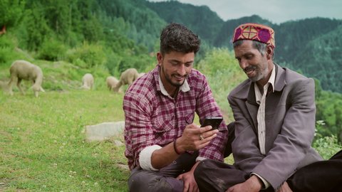 Smiling Indian elderly male shepherd with his young son sitting together using a Mobile phone with grazing sheep in a rural area of remote greenery mountainous region. Entertainment and technology 