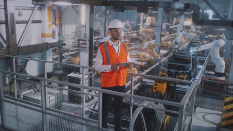 Pan around high angle view of male employee in uniform and hardhat browsing tablet against automatic machines on modern vehicle factory