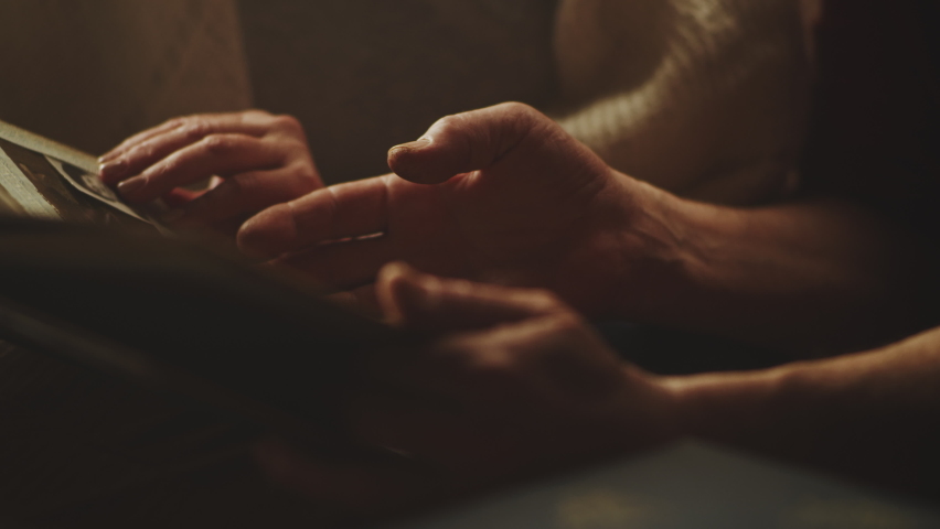 Crop senior husband and wife holding hands and flipping pages of photo album while recollecting old memories in weekend at home