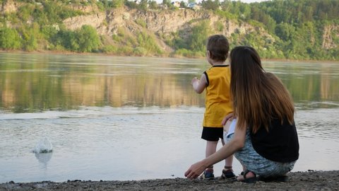 A cute girl and a child on the river bank throw pebbles into the water