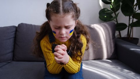 Hopeful little girl kid keep hands in prayer hope sitting on sofa at home, child with flag of ukraine on face. Children against war. Russia's invasion of Ukraine, request for world community's help