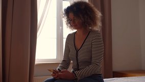 Ukrainian woman typing message on mobile phone while sitting in dark bedroom alone. Curly young white female texting online on social media messenger with modern smartphone
