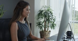 A corporate Asian woman seated near a large office window calmly types and smiles working at a desktop computer. 35mm Medium Closeup Tripod Shot 4K.